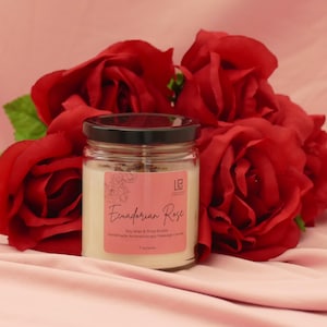 May include: A clear glass jar candle with a black lid and a pink label reading Ecuadorian Rose, Soy Wax & Shea Butter, Handmade Aromatherapy Massage Candle, 7 ounces. The candle is surrounded by a cluster of deep red artificial roses.