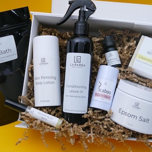 May include: A white gift box filled with self-care products, including a black bag labeled "Sitz Bath", a bottle of "Conditioning Leave In" spray, and a jar of "Epsom Salt". Other items include a lip balm and a bar of soap.