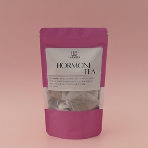 Pu&ograve; includere: Un sacchetto magenta di "Hormone Tea" con un'etichetta bianca. L'etichetta include il marchio "La Parea Wellness" e un elenco di ingredienti. Il contenuto delle bustine di t&egrave; &egrave; visibile attraverso una finestra trasparente.
