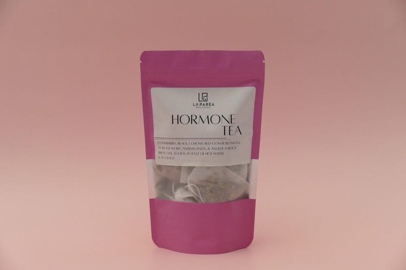Pu&ograve; includere: Un sacchetto magenta di "Hormone Tea" con un'etichetta bianca. L'etichetta include il marchio "La Parea Wellness" e un elenco di ingredienti. Il contenuto delle bustine di t&egrave; &egrave; visibile attraverso una finestra trasparente.