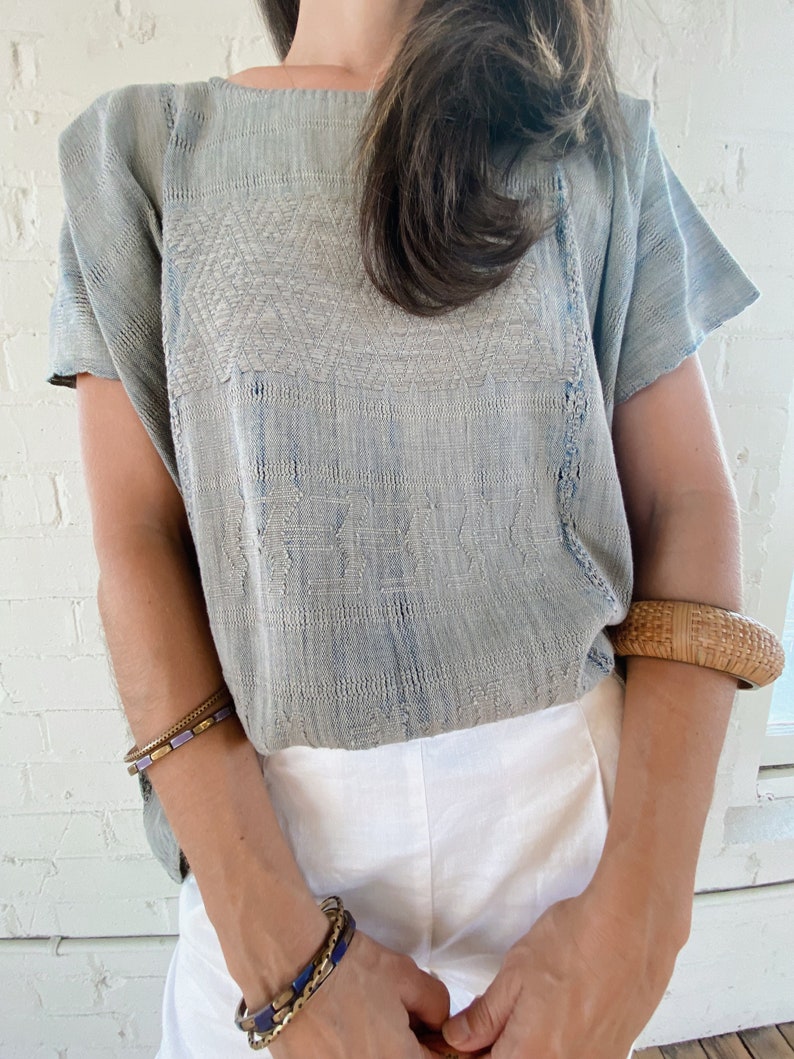 May include: A light blue embroidered cotton blouse with short sleeves. The blouse has a relaxed fit and a round neckline. The embroidery is a geometric pattern in a lighter shade of blue.