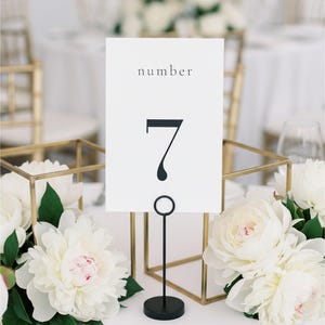 以下が含まれることがあります： 「number」の文字と黒い数字の7が書かれた白いテーブルナンバーカード。カードは黒い金属製のスタンドで支えられています。テーブルセッティングには白い花と金の幾何学的な装飾が含まれています。