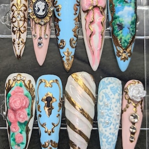 May include: Ten long, pointed, artificial fingernails with various designs. The designs include a cameo, a bow, a landscape, roses, a keyhole, a swirl, and a floral pattern. The nails are painted in shades of pink, blue, and white with gold accents.