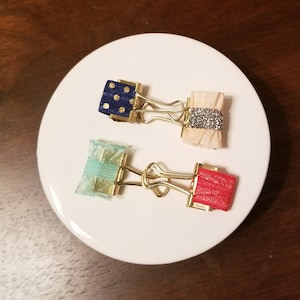 Puede incluir: Tres clips de carpeta dorados con lazos decorativos. Los lazos son de color azul con lunares blancos, beige con purpurina plateada y rojo.