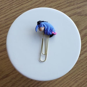 May include: A gold paper clip with a blue and pink Eeyore character attached to the top.