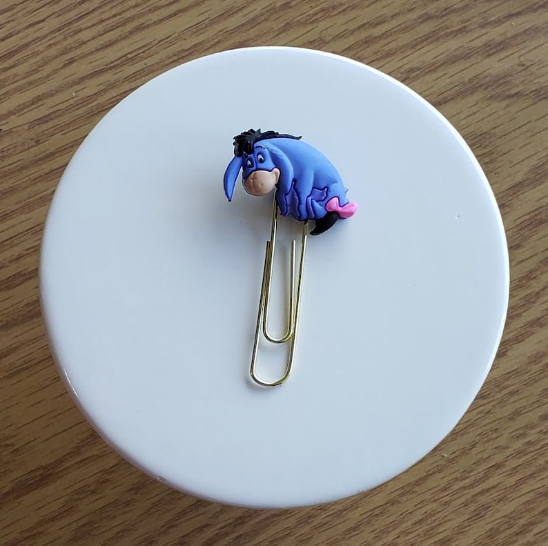 May include: A gold paper clip with a blue and pink Eeyore character attached to the top.
