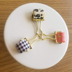 May include: Three gold binder clips with patterned fabric covers. The covers are black and white gingham, leopard print, and red with a gold flamingo silhouette.