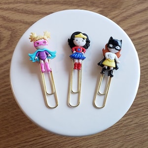 May include: Three gold paper clips with colorful superhero character toppers. The characters are a girl with a purple suit and blue cape, a girl with a red and blue suit and a star on her chest, and a girl with a yellow suit and a black mask.
