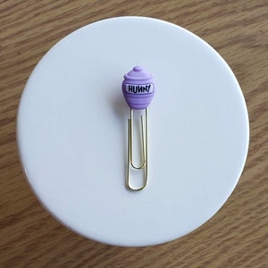 May include: Purple honey pot shaped paper clip with the word "Hunny" on it. The paper clip is gold.