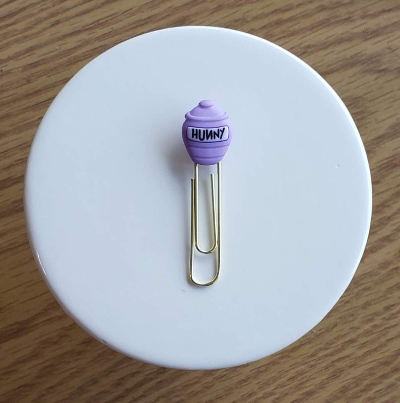 May include: Purple honey pot shaped paper clip with the word "Hunny" on it. The paper clip is gold.