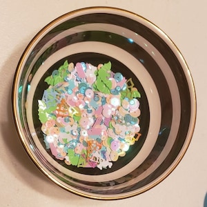 May include: A decorative bowl with black and white stripes and a gold rim. The bowl is filled with colorful sequins and confetti in pastel shades of pink, blue, green, and orange. The sequins are in various shapes, including flowers and leaves.