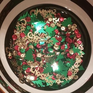 Op de afbeelding: Een kom gevuld met groene en gouden kerstboomvormige confetti en rode en gouden ronde confetti. De confetti spelt "Merry Christmas".