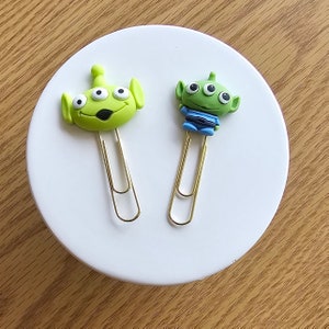 May include: Two gold paper clips with green alien character toppers. One alien is standing with its arms outstretched and the other is laying down with its eyes closed.