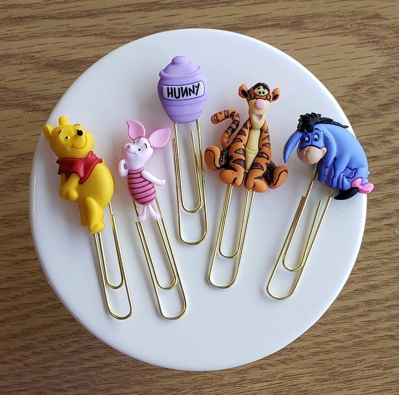 May include: Five gold paper clips with different Winnie the Pooh character toppers. The toppers include Winnie the Pooh, Piglet, Tigger, Eeyore, and a jar of honey that says "Hunny".
