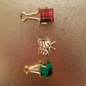 May include: Three gold binder clips decorated with festive holiday-themed patterned fabric. The top clip has a red and black plaid pattern, the middle clip has a red and green striped pattern, and the bottom clip has a green pattern with small Christmas trees.