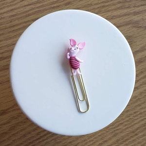 May include: A gold paper clip with a pink pig character attached to the top. The pig has a pink body with red stripes and a pink snout.