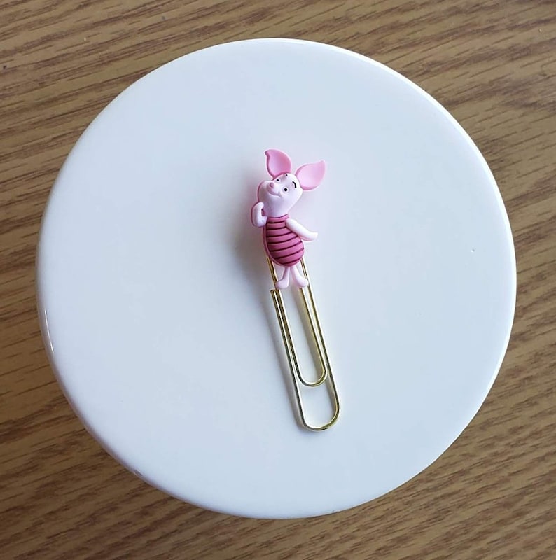 May include: A gold paper clip with a pink pig character attached to the top. The pig has a pink body with red stripes and a pink snout.