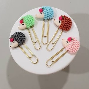 May include: Five decorative paper clips with hedgehog designs. Each clip features a different color hedgehog: green, blue, brown, pink, and gray. The hedgehogs have a white face, a small pink flower, and gold-colored metal clips.