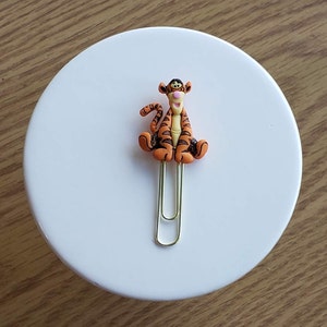 May include: A gold paper clip with an orange and black Tigger character attached. The character is sitting with its legs crossed and has a pink nose.