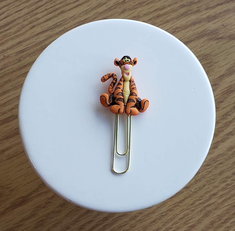 May include: A gold paper clip with an orange and black Tigger character attached. The character is sitting with its legs crossed and has a pink nose.