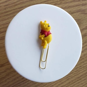 May include: A gold paperclip with a yellow Winnie the Pooh character attached to the top. The character is standing upright with its arms crossed.
