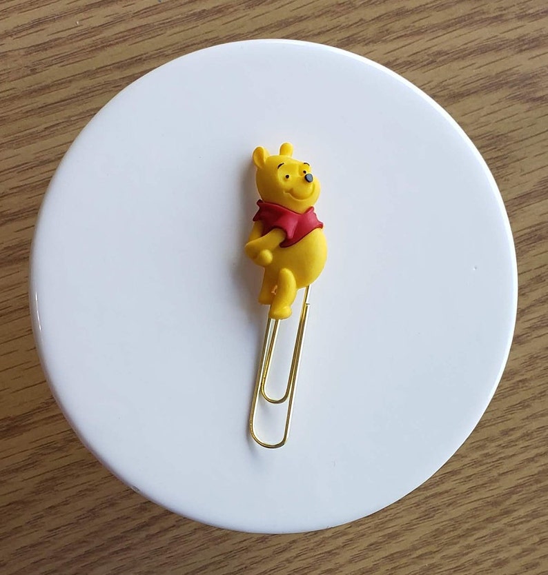 May include: A gold paperclip with a yellow Winnie the Pooh character attached to the top. The character is standing upright with its arms crossed.
