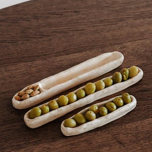 Könnte beinhalten: Drei cremefarbene Keramik-Servierschalen auf einer Holzoberfläche. Die langen, schmalen Schalen sind mit grünen Oliven und Nüssen gefüllt. Geeignet für Vorspeisen oder Snacks.
