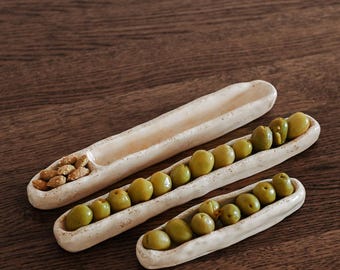 May include: Three off-white ceramic serving dishes on a wooden surface. The dishes are long and narrow, filled with green olives and nuts. Ideal for presenting appetizers or snacks.