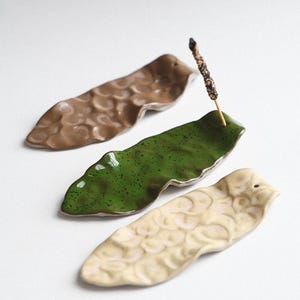 May include: Three ceramic incense holders in shades of brown, green, and cream. The holders are leaf-shaped with textured surfaces. One green holder holds a lit incense stick. The holders are arranged on a white surface.