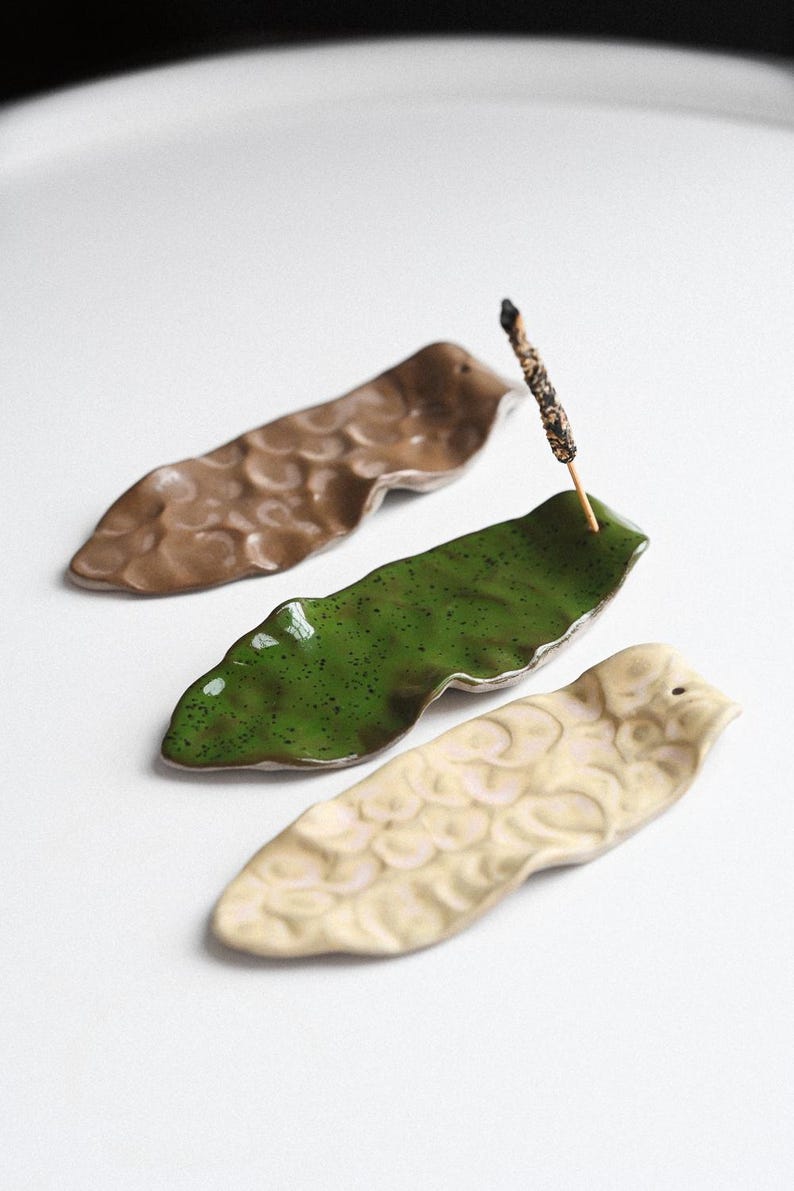 May include: Three ceramic incense holders in shades of brown, green, and cream. The holders are leaf-shaped with textured surfaces. One green holder holds a lit incense stick. The holders are arranged on a white surface.