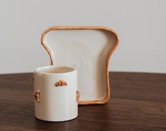 May include: A ceramic bread-themed set featuring a mug with croissant accents and a bread-shaped plate. The mug and plate are off-white with brown trim, set on a dark wooden surface. The set is designed to resemble a slice of bread.