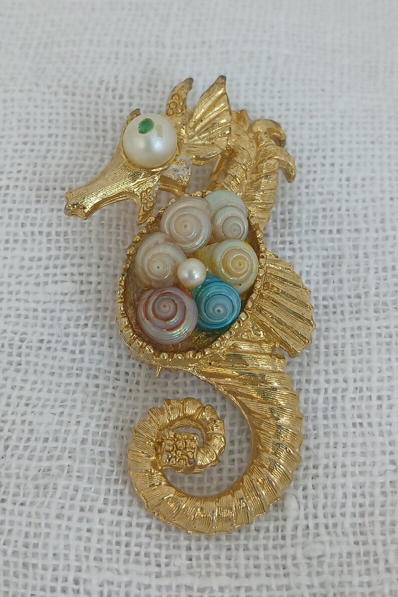 May include: A gold-tone seahorse brooch with a pearl eye and a nest of colorful seashells.
