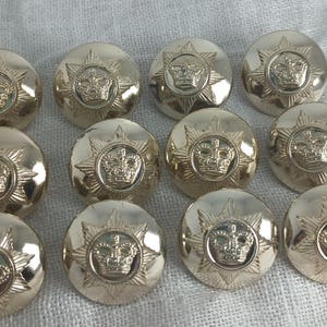 May include: A collection of twelve gold-colored buttons. Each button features a crown design at the center, surrounded by a starburst pattern. The buttons are round and have a shiny, metallic finish. They are arranged on a white, textured surface.
