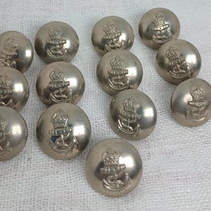 Puede incluir: Colección de botones metálicos plateados. Cada botón presenta una corona sobre un diseño de ancla. Los botones son redondos y tienen una forma ligeramente abovedada. Están dispuestos sobre una superficie blanca y texturizada.