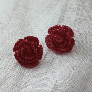 May include: Two handmade, dark red crochet flower earrings. The earrings are in the shape of roses, with multiple layers of petals. They are set against a white, textured background.