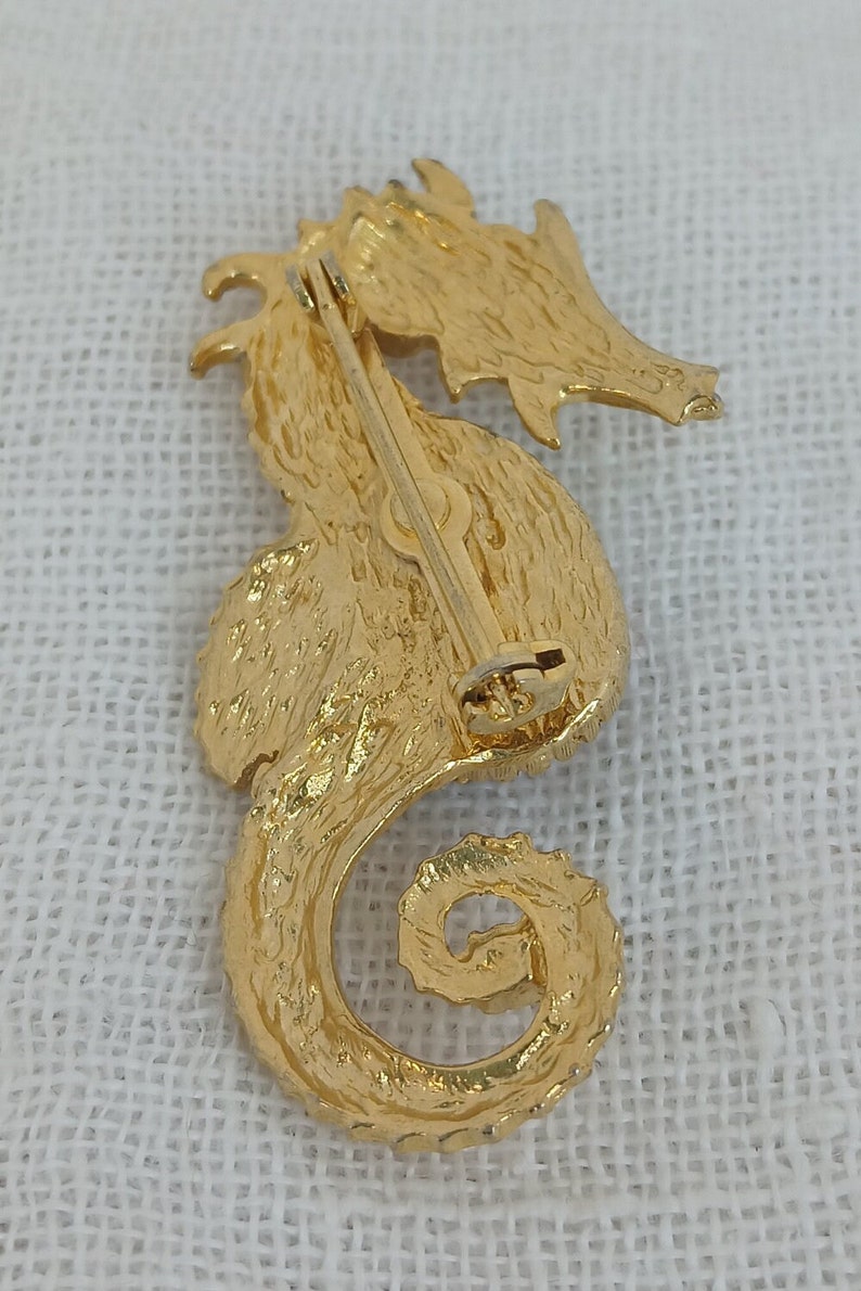 May include: Gold-toned seahorse brooch with a textured finish. The brooch has a pin and catch closure on the back.