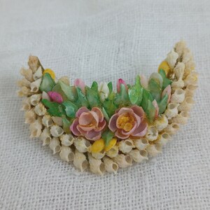 以下が含まれることがあります： 貝殻で作られた三日月型のブローチで、花束のようにペイントされています。ブローチには、ピンクと白の花と緑の葉が描かれており、すべて貝殻で作られています。