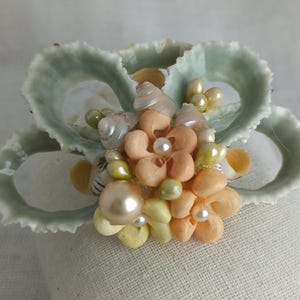 May include: A handcrafted seashell and pearl floral arrangement. The arrangement features a cluster of peach, yellow, and white seashells and pearls, set against a backdrop of pale green scallop shells. A unique, decorative item.