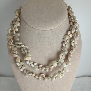 May include: A layered necklace made of small, natural seashells in shades of cream, beige, and light brown. The necklace is displayed on a beige mannequin bust, showcasing its intricate design and texture. The shells vary in size and shape.