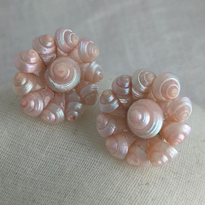 May include: Two seashell-shaped earrings. Each earring is composed of multiple small, iridescent, pale pink seashells clustered together. The shells have a spiral design and a pearlescent sheen, creating a shimmering effect. The earrings are set against a neutral background.