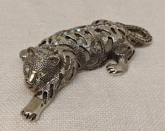 May include: A silver-toned tiger brooch with clear iridescent rhinestones. The tiger is in a crouching position with its head turned to the side.