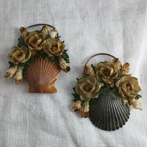 May include: Two seashell-shaped decorative pieces. Each features a basket of flowers made of seashells and faux greenery. The flowers are a mix of tan and gold, with green leaves. The shell baskets are tan and dark gray.