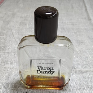 Może przedstawiać: Przezroczysta szklana butelka z wodą kolońską Varon Dandy International. Butelka ma czarną zakrętkę i brązowy płyn w środku.