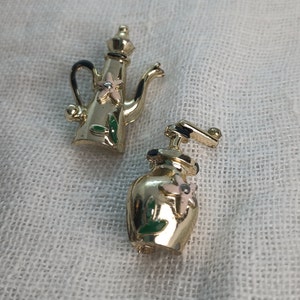 May include: Two gold-tone metal brooches in the shape of a coffee pot and a coffee grinder. Both brooches have green leaves and pink flowers painted on them.