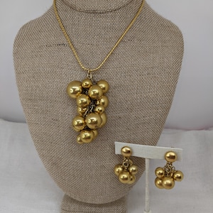 Può includere: Un set di collana e orecchini in tono oro con un gruppo di sfere rotonde. La collana è in stile catena a maglie e gli orecchini sono a goccia.