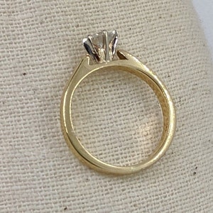 K&ouml;nnte beinhalten: Ein goldener Verlobungsring mit einem einzelnen runden Diamanten in einer Krappenfassung.