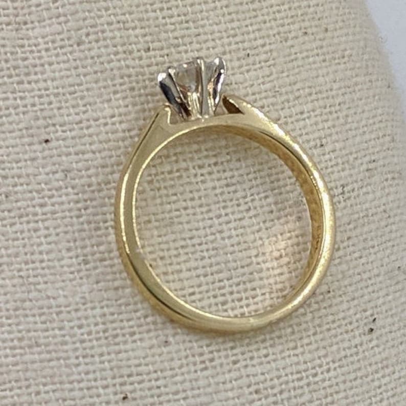 K&ouml;nnte beinhalten: Ein goldener Verlobungsring mit einem einzelnen runden Diamanten in einer Krappenfassung.