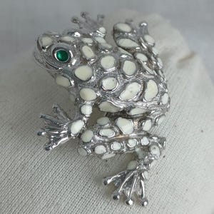 May include: A silver frog brooch with white enamel spots and a green eye. The frog's design is detailed, with textured skin and articulated legs. A unique accessory.