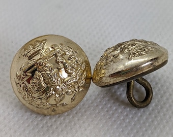 Könnte beinhalten: Zwei goldfarbene Metallknöpfe mit aufwendigen Designs. Ein Knopf zeigt ein detailliertes Blumenmuster, während der andere ein abstrakteres Design hat.