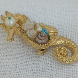 May include: A gold-tone seahorse brooch with a pearl and green stone eye. The seahorse is holding a cluster of iridescent seashells.